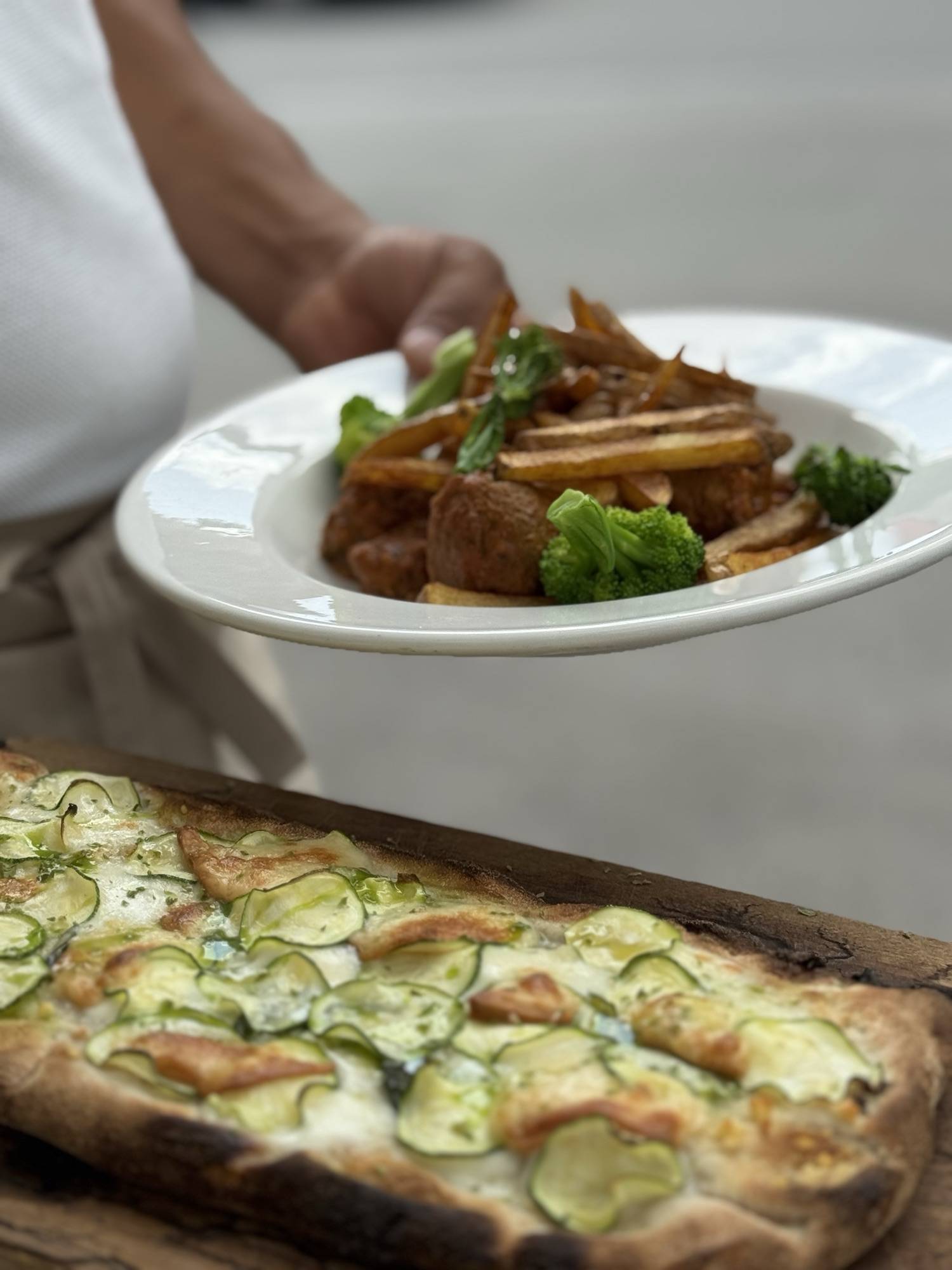 Courgette pinsa fresh from the oven on a wooden board, a main course being served behind it